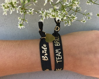 Op de afbeelding: Twee zwarte elastische polsbandjes met gouden letters. Een polsbandje zegt "Bride" en het andere zegt "Team Bride".