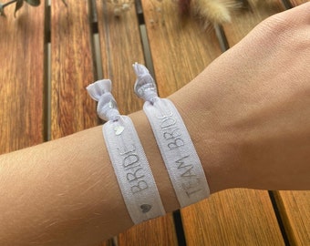 Op de afbeelding: Twee witte elastieken armbandjes met zilveren opschrift. Eén armbandje zegt "Team Bride" en het andere zegt "Bride" met een klein hartje aan elke kant van de tekst.