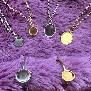 May include: A collection of necklaces and rings with settings for small cabochons. The necklaces have delicate chains in gold and silver tones, with round and oval settings. The rings have similar settings in gold and silver.