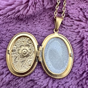 K&ouml;nnte beinhalten: Ge&ouml;ffneter goldfarbener Medaillon-Anh&auml;nger an einer Goldkette. Das Medaillon ist oval und hat auf einer Seite ein eingraviertes Blumendesign. Die andere Seite ist leer. Der Hintergrund ist ein verschwommener lila Stoff.