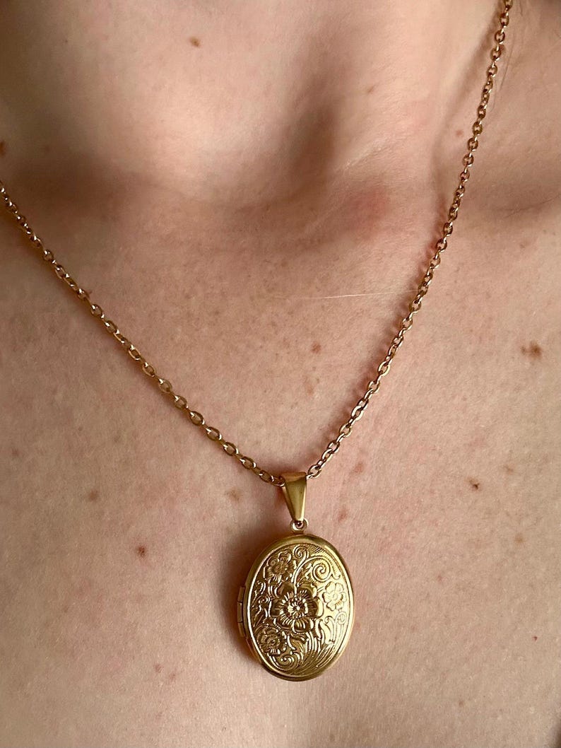K&ouml;nnte beinhalten: Goldfarbene ovale Medaillon-Anh&auml;nger-Halskette. Das Medaillon hat ein florales Design und h&auml;ngt an einer zarten Kette. Ein klassisches Schmuckst&uuml;ck.