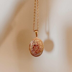 K&ouml;nnte beinhalten: Eine goldfarbene ovale Medaillon-Halskette. Das Medaillon hat ein gepr&auml;gtes Blumendesign und h&auml;ngt an einer zarten Kette. Der Anh&auml;nger ist etwa 3,2 cm lang.