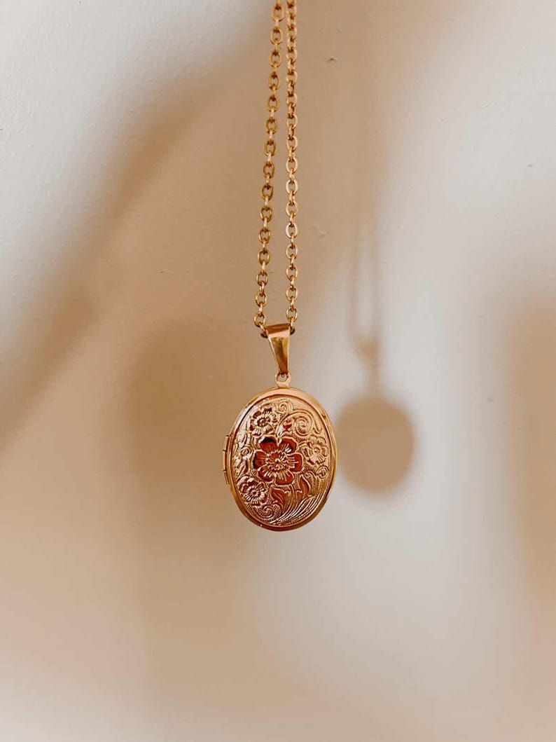 K&ouml;nnte beinhalten: Eine goldfarbene ovale Medaillon-Halskette. Das Medaillon hat ein gepr&auml;gtes Blumendesign und h&auml;ngt an einer zarten Kette. Der Anh&auml;nger ist etwa 3,2 cm lang.