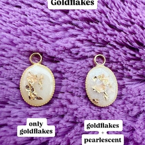 May include: Two oval pendants with gold flakes and a gold-colored border. One pendant is labeled "only goldflakes", and the other is labeled "goldflakes + pearlescent". The background is a textured purple fabric, and the text "Optional Add-On: Goldflakes" is at the top.
