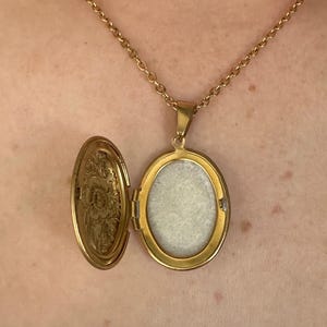 K&ouml;nnte beinhalten: Goldfarbene Medaillon-Halskette mit ovalem Anh&auml;nger, ge&ouml;ffnet, um einen hellen Innenraum zu zeigen. Das Medaillon h&auml;ngt an einer zarten Kette. Die Innenseite des Medaillons hat ein Blumenmuster.