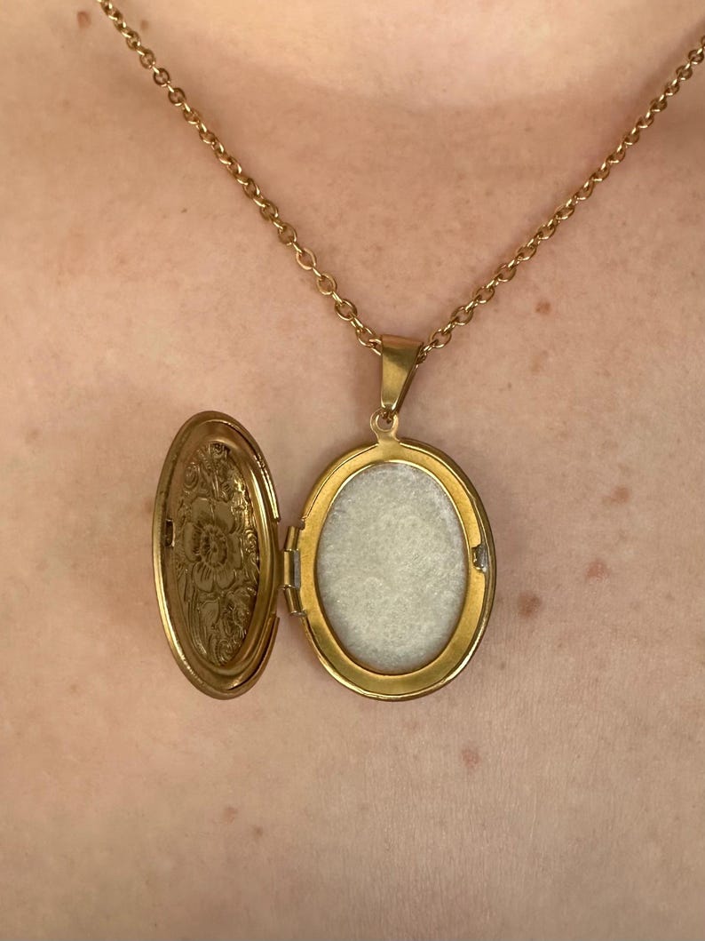 K&ouml;nnte beinhalten: Goldfarbene Medaillon-Halskette mit ovalem Anh&auml;nger, ge&ouml;ffnet, um einen hellen Innenraum zu zeigen. Das Medaillon h&auml;ngt an einer zarten Kette. Die Innenseite des Medaillons hat ein Blumenmuster.