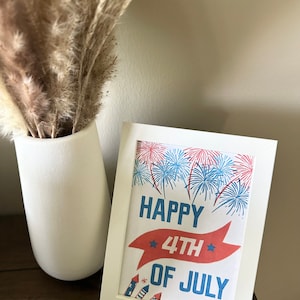 以下が含まれることがあります： 赤、白、青の打ち上げ花火と「Happy 4th of July」という赤いバナーのテキストが書かれた白い額縁入りプリントです。