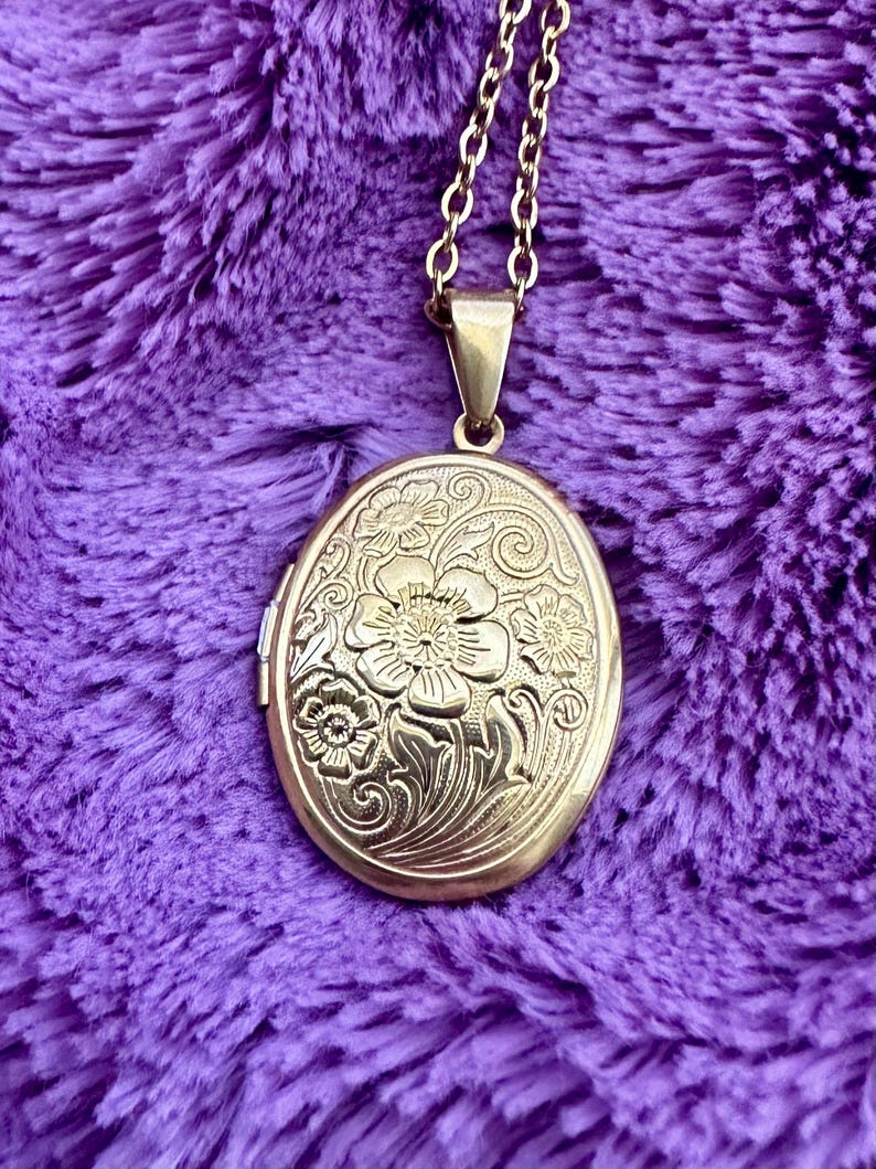 K&ouml;nnte beinhalten: Goldfarbene ovale Medaillon-Halskette mit floraler Gravur. Das Medaillon h&auml;ngt an einer filigranen Kette. Der Hintergrund ist ein strukturierter, violetter Stoff.