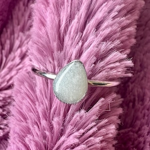 Puede incluir: Una pulsera plateada con una piedra brillante en forma de lágrima, de color claro. La piedra está engastada en un bisel plateado y se asienta sobre una fina banda plateada. La pulsera se muestra sobre una superficie morada y esponjosa.