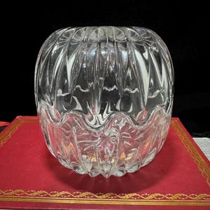 May include: A clear, round glass bowl with a ribbed exterior design. The bowl sits on a red book with gold trim, against a black background. The bowl's design features vertical and curved lines, creating a textured appearance.