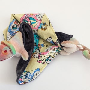 May include: A colorful plush toy chameleon with a rainbow pattern and a black and white patterned fabric blanket. The blanket has a cartoon design with people and umbrellas.