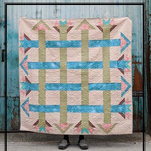 May include: A beige quilt with a geometric pattern of green, blue, and pink arrows. The quilt is hanging on a blue wooden wall.