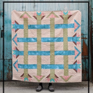 May include: A beige quilt with a geometric pattern of green, blue, and pink arrows. The quilt is hanging on a blue wooden wall.