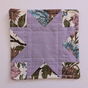 May include: Square quilted coaster with a central lavender star design. The star is surrounded by floral patterned fabric in shades of pink, blue, green, and brown. The coaster is likely made of cotton or a similar fabric.