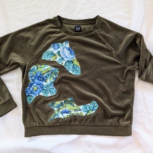 May include: A dark green velour sweatshirt with a floral pattern on the left side. The floral pattern is made up of blue, green, and white flowers. The sweatshirt has a crew neck and long sleeves.