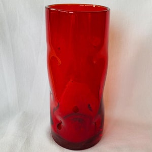 May include: A tall, red glass vase with a unique, irregular shape. The vase has a slightly textured surface and a rounded bottom.