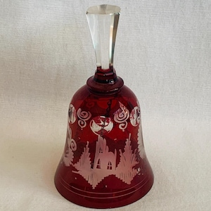 May include: A ruby red glass bell with a clear, faceted handle. The bell features etched designs of a winter scene with trees and a building. A decorative item for display.