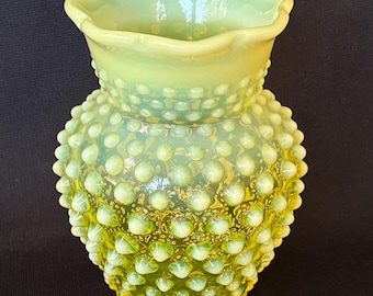 May include: A translucent lime green glass vase featuring a hobnail texture with raised bumps across the body. The vase has a wide, scalloped rim and a rounded, bulbous shape that tapers toward the base, showcasing a vibrant, light-catching finish.