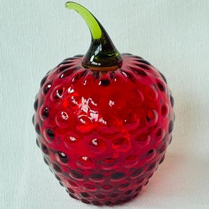May include: A red glass decorative bottle shaped like a strawberry. The bottle has a textured surface with small, round bumps and a green stem. The glass is translucent, allowing light to pass through.
