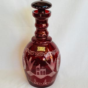 May include: A ruby red glass decanter with a matching stopper. The decanter features etched landscape scenes with buildings and trees. A small sticker is visible on the front. The decanter is a decorative item.