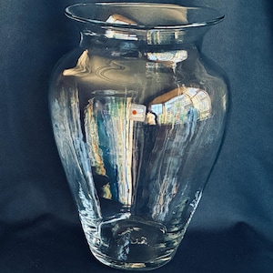 May include: A clear glass vase with a wide, rounded body and a flared rim. The vase reflects the surrounding environment, creating distorted images within its transparent form. The vase sits against a dark blue background.