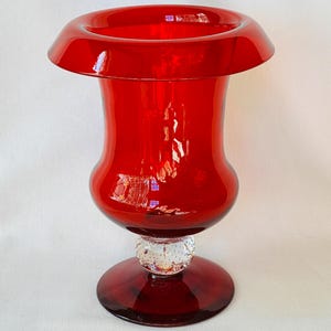 May include: A vibrant red glass vase with a flared top and a bulbous body. The vase sits on a clear, textured base and a circular foot. The overall height is approximately 8 inches.