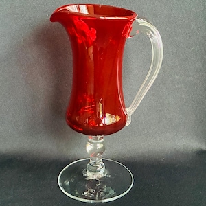 May include: A tall, vibrant red glass pitcher featuring a smooth, flared body with a distinct pouring spout. The vessel is supported by a clear glass pedestal base and includes a curved, transparent glass handle attached to the side.
