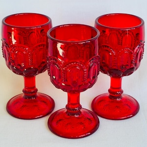 May include: Three ruby red goblets with ornate, embossed detailing. The stemware features a rounded bowl, a short stem, and a circular base. The glasses are made of transparent glass and are arranged on a white surface. The goblets are likely vintage.