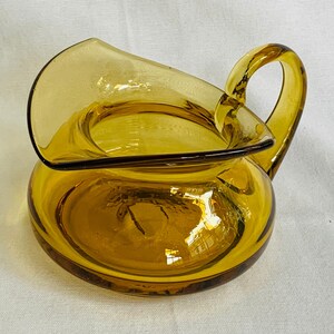 May include: Amber glass pitcher with a unique, curved spout and a handle. The pitcher has a rounded base and a smooth, glossy finish.