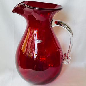 May include: A deep red glass pitcher with a bulbous body, a wide flared spout, and a clear glass handle. The pitcher features a smooth, glossy finish and a small, clear glass decorative knot detail at the base of the handle, reflecting light.