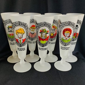 May include: A set of eight frosted glass champagne flutes. Each flute features a different cartoon character portrait within a black and white ornate frame. The names "Papa", "Mama", "Uncle Bert", "Victoria", "Aunt Aggie", "Rupert", and "Auntie" are printed below the portraits.