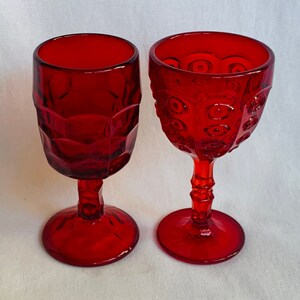 May include: Two red glass goblets with different patterns. One goblet has a textured, diamond-like pattern, while the other has a raised dot pattern.