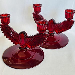 May include: A pair of ruby red glass candle holders, each featuring a circular base, a central vertical fin-like element, and two side arms that extend outward to support individual candle cups with a scalloped, serrated rim design.