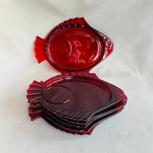 May include: Set of four red glass fish-shaped plates. The plates are stacked on top of each other. The plates have a raised rim and a small indentation in the center.