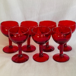 May include: Set of seven ruby red wine glasses. Each glass features a rounded bowl, a slender stem, and a circular base. The glasses are arranged on a white surface, showcasing their vibrant color and classic design. Ideal for serving wine.