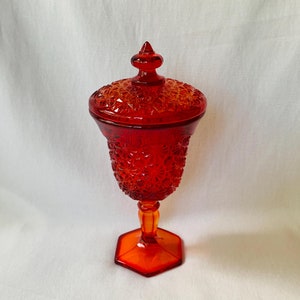 May include: A red glass candy dish with a lid. The dish has a pedestal base and a detailed, patterned design.