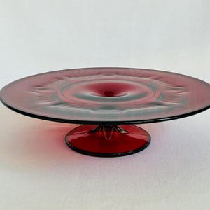 May include: A ruby red glass cake stand with a circular design on the plate. The stand has a pedestal base and a wide, flat top. The glass has a glossy finish, and the stand is likely used for displaying desserts or other food items.