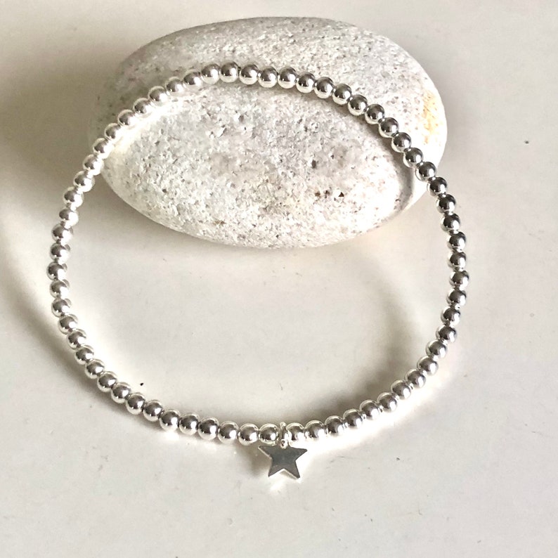 May include: A silver beaded bracelet with a small star charm. The bracelet is made of small, round, silver beads strung together. The star charm hangs from the center of the bracelet. The bracelet is displayed on a white surface.