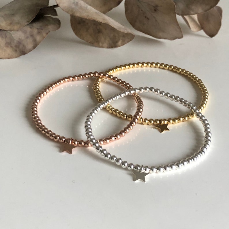 May include: Three beaded bracelets in rose gold, gold, and silver. Each bracelet features small, round beads and a star charm. The bracelets are displayed on a white surface, with a blurred background of dried leaves.