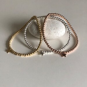May include: Three beaded bracelets in gold, silver, and rose gold, each with a small star charm. The bracelets are strung with small, round beads and rest on a white surface. The bracelets are a delicate jewelry item.