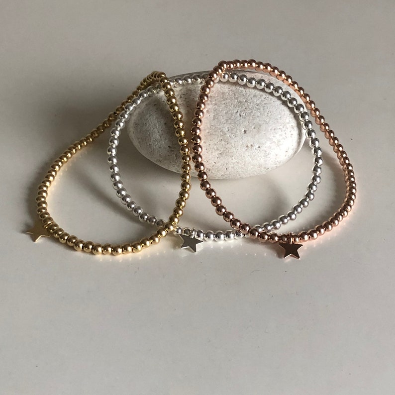 May include: Three beaded bracelets in gold, silver, and rose gold, each with a small star charm. The bracelets are strung with small, round beads and rest on a white surface. The bracelets are a delicate jewelry item.