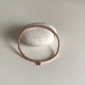 May include: Rose gold beaded bracelet with a star charm. The bracelet is made of small, round beads strung together, forming a flexible band. A small star charm hangs from the bracelet. The bracelet is displayed on a white stone.