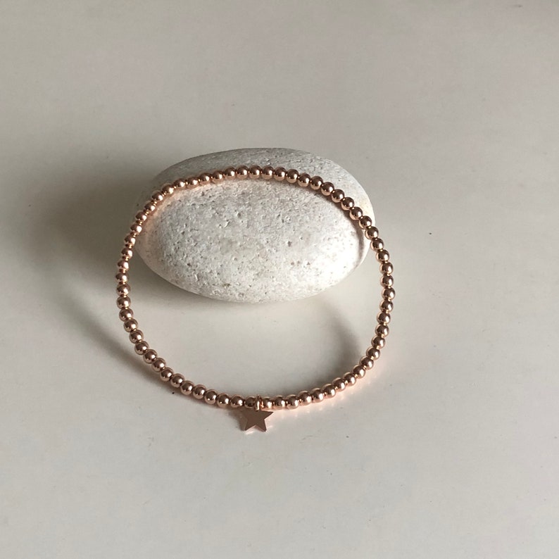 May include: Rose gold beaded bracelet with a star charm. The bracelet is made of small, round beads strung together, forming a flexible band. A small star charm hangs from the bracelet. The bracelet is displayed on a white stone.