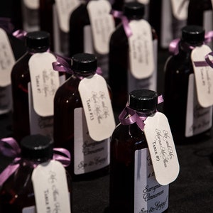 May include: A collection of small, dark glass bottles with black caps, each adorned with a white tag and a purple ribbon. The tags feature elegant script and the words "Mr. and Mrs" and "Table #". The bottles contain a reddish-brown liquid.