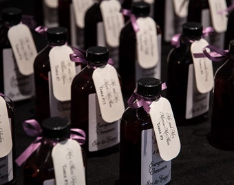May include: Several small, dark glass bottles with black caps, each decorated with a white tag and a purple ribbon. The tags display elegant script and the words "Mr. and Mrs" and "Table #". The bottles contain a reddish-brown liquid.