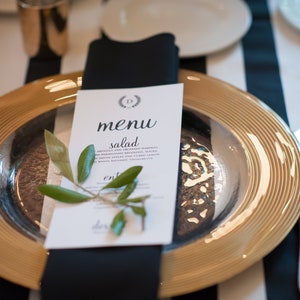 May include: A formal table setting featuring a gold-rimmed charger plate with a silver interior. A black napkin holds a menu card with the word "menu" and a sprig of greenery. The background has black and white stripes.