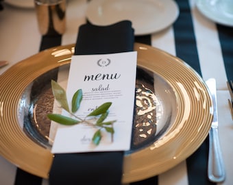 May include: A formal table setting with a gold-edged charger plate and a silver interior. A black napkin holds a menu card with the word "menu" and a sprig of greenery. The background has black and white stripes.