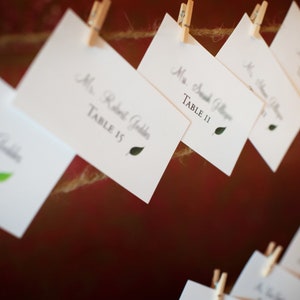 May include: White place cards with names and table numbers are clipped to a twine line with small wooden clothespins. Each card has a green leaf graphic. The background is a blurred, dark red.