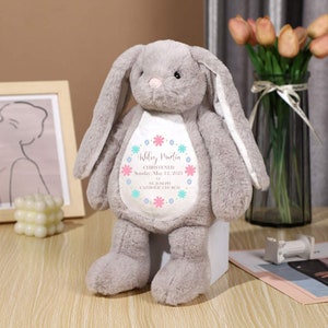 Puede incluir: Un conejito de peluche gris con orejas largas y una nariz rosa. El conejito tiene un parche blanco en el pecho con el texto "Ashley Martin CHRISTENED Sunday, May 12, 2023 ST. JOSEPH CATHOLIC CHURCH" rodeado por un diseño floral.