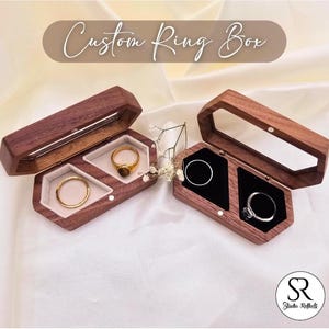 Può includere: Due scatole per anelli in legno con coperchio trasparente. Le scatole sono in legno scuro e hanno una forma esagonale. Ogni scatola ha due scomparti per anelli. Le scatole sono perfette per riporre ed esporre anelli.